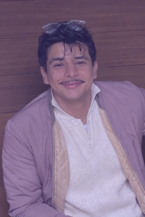 Portrait of a smiling young man with dark hair, a mustache, and glasses on his head, wearing a light purple jacket against a wood-paneled wall